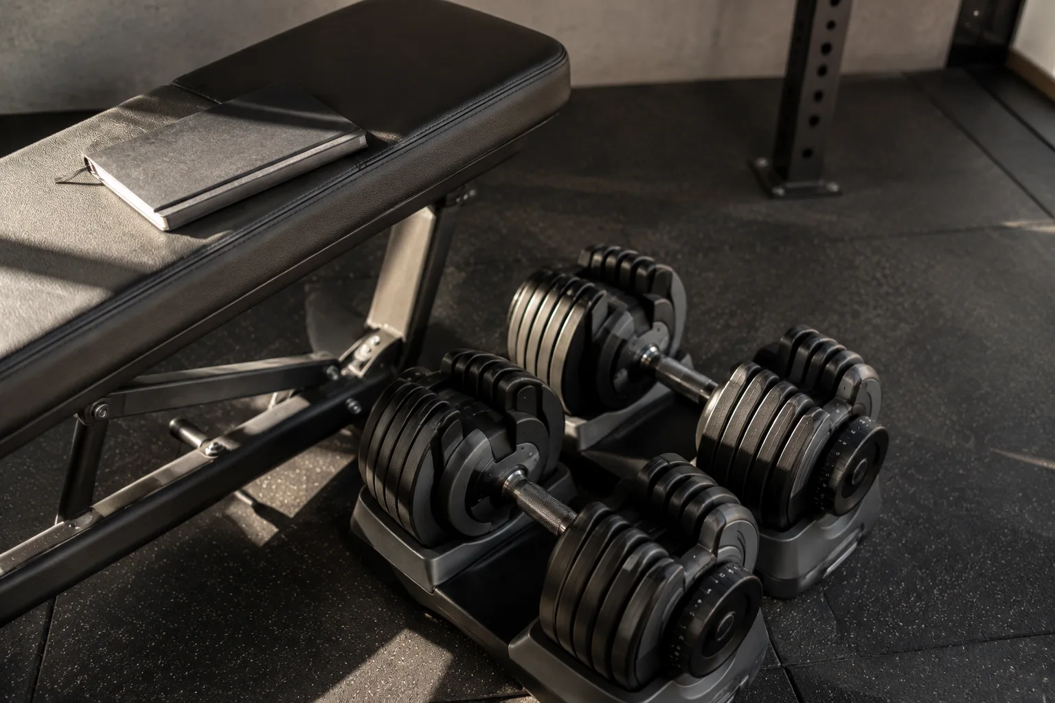 A pair of adjustable dumbbells beside a gym bench with a workout notebook